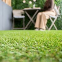 Ein junges Paar steht barfuß auf einem Balkon mit Kunstrasen und hält jeweils eine Tasse in der Hand. Beide lächeln, die Frau lehnt sich entspannt an den Mann. Um sie herum stehen Blumentöpfe mit bunten Blumen und eine Pflanze. Oben im Bild steht der Text „Mit GUT zertifiziert“. Rechts daneben sind ein Siegel mit Baum-Logo und der Schriftzug „Made in EU“ zu sehen. Im Hintergrund sind moderne Wohnhäuser sichtbar.