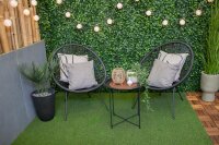 Dachterrasse mit Kunstrasen, weißen Pflanzkübeln voller Kräuter und Gräsern, einem modernen Holztürelement mit Glas und Jalousie, darüber hängender Efeupflanze, daneben vier hohe weiße Dekovasen auf Kieselsteinen vor einer weißen Wand mit schrägem Dach und kleinem Türchen. Im Hintergrund ist ein heller Wohnraum mit weißen Stühlen sichtbar, der Himmel ist leicht bewölkt.