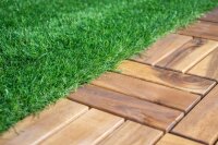 Moderner, ebenerdiger Terrassenbereich eines Hauses mit hellen, glatten Wänden, großen Glasfronten und Holzboden. Im Vordergrund steht ein kleiner Baum auf grünem Rasen, auf der Terrasse stehen dunkle Rattanmöbel mit roten Polstern. Links ist eine dekorative Holzsäulenverkleidung, im Hintergrund sind Rasen und Bäume sichtbar, der Himmel ist bewölkt.